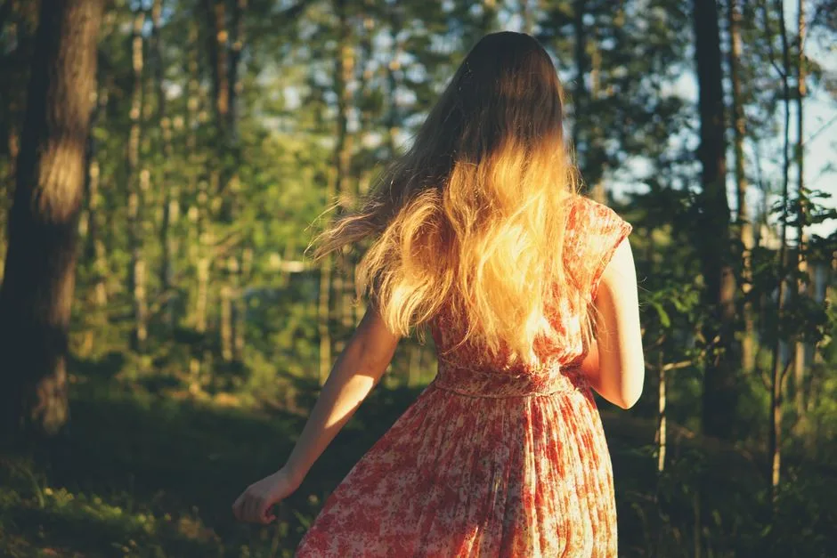 A woman in a floral dress walks through a sunlit forest. Bright back view.