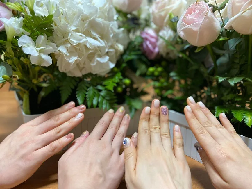 Group of hands with varied nail art surrounded by lush flower arrangements in pastel tones.