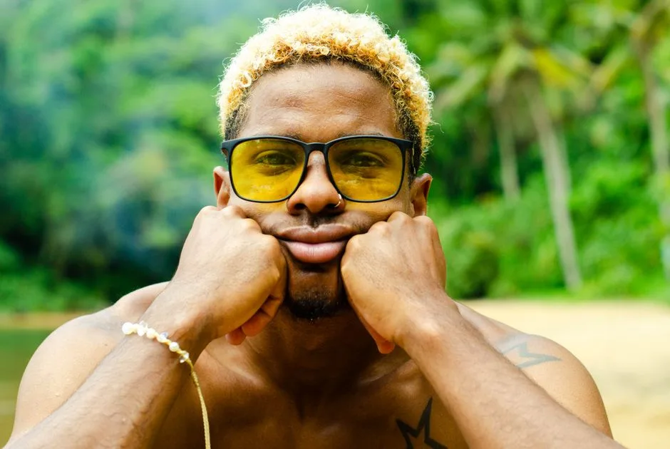 Close-up of a young man with glasses and blonde hair, resting on hands, in a tropical setting.