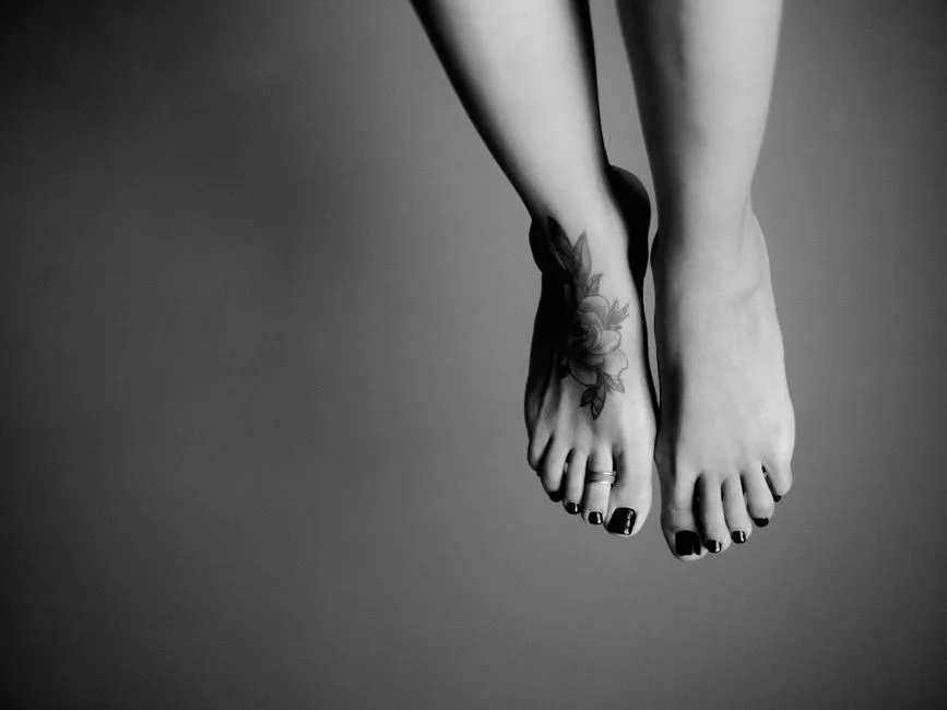 Black and white artistic photo highlighting tattooed feet with dark nail polish.