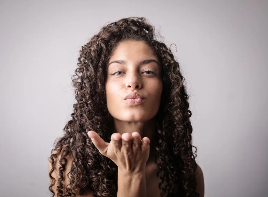 Elegant young woman with curly hair sending a playful kiss in studio. Perfect for beauty and lifestyle themes.