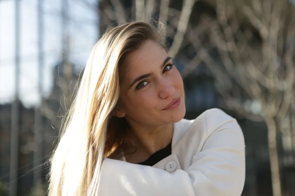 A young woman smiling in a sunny outdoor setting, showcasing blonde hair and casual style.