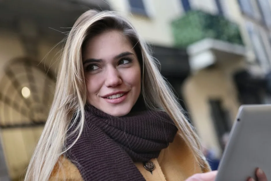 A cheerful young woman with blond hair and a scarf smiles while standing outdoors with her tablet.