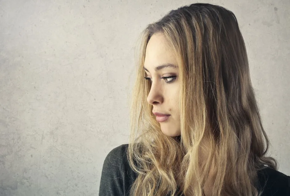 A pensive young woman with long blonde hair looking to the side against a neutral background.