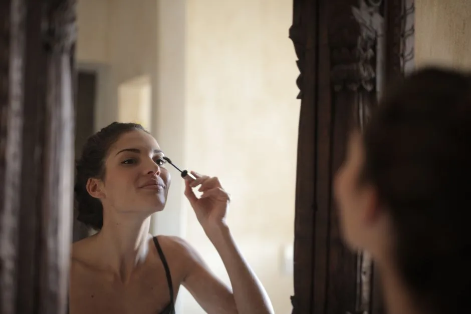 A woman applies mascara while looking in a stylish wooden mirror indoors.
