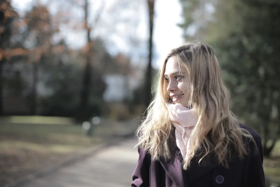 A young blonde woman smiling as she stands outside on a sunny winter day, wearing a violet coat and scarf.