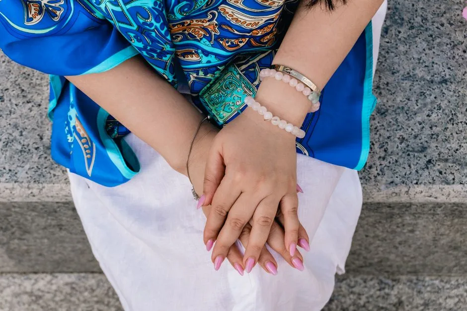 Close-up of a woman's hands with pink nails, wearing bracelets and a blue patterned dress.