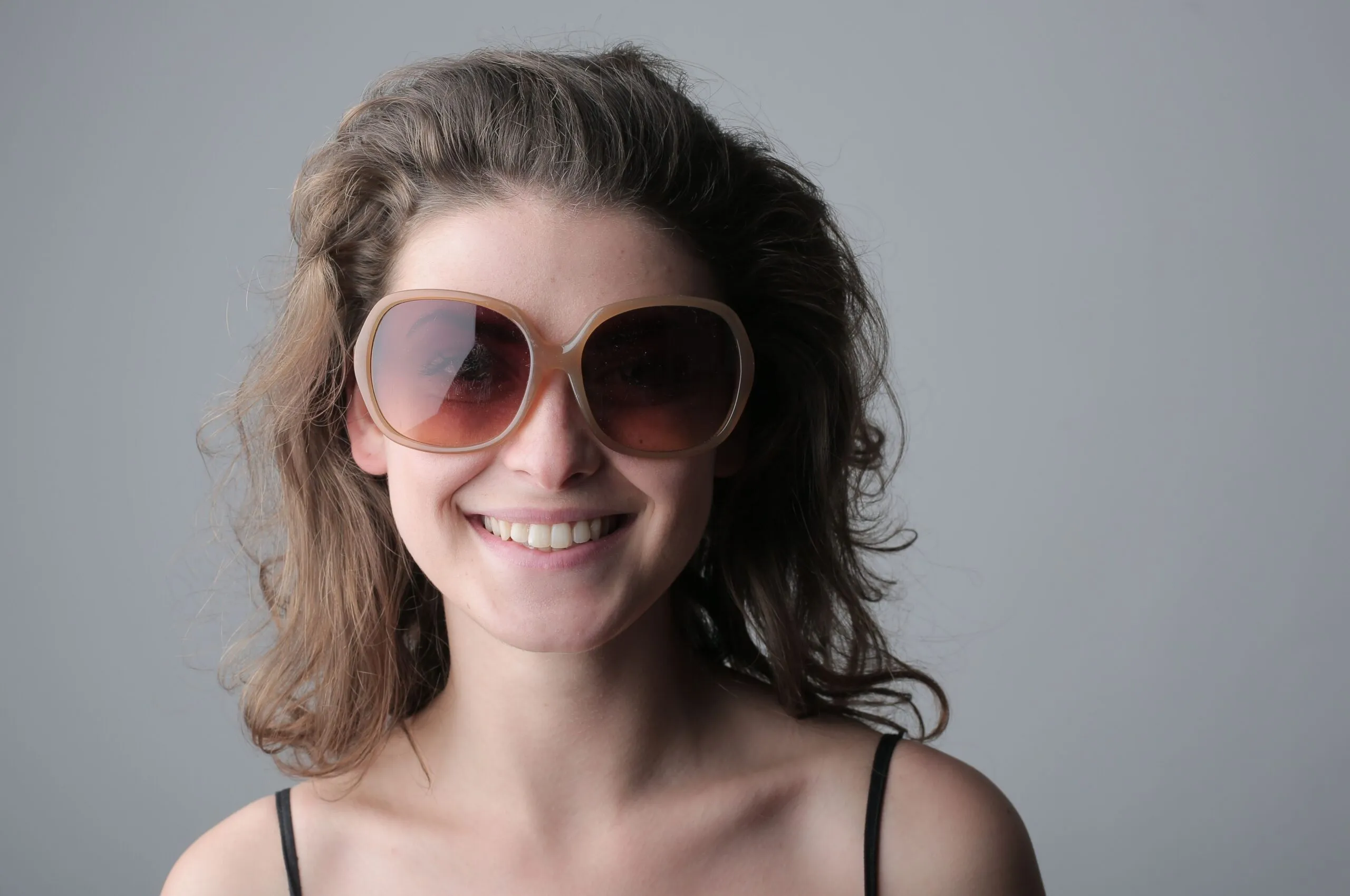 Portrait of a smiling woman with wavy hair and sunglasses against a neutral background.