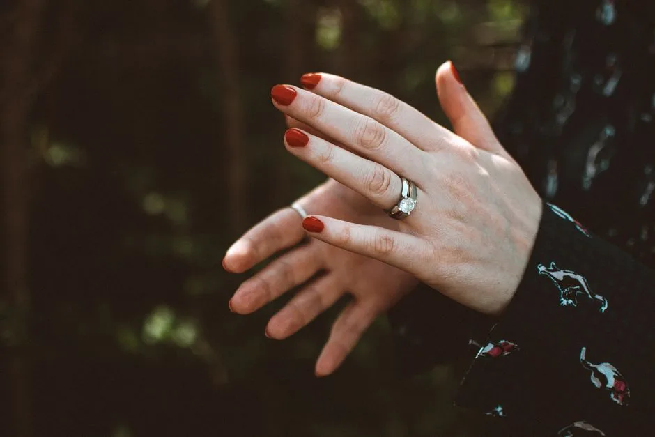 Close-up of hands with red nails showcasing a diamond ring outdoors, symbolizing luxury and elegance.