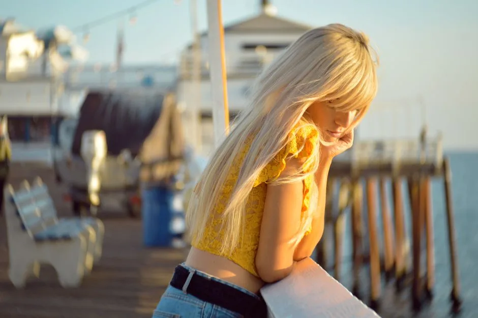 A blonde woman with long hair leans on a railing at Malibu Pier during sunset, deep in thought.