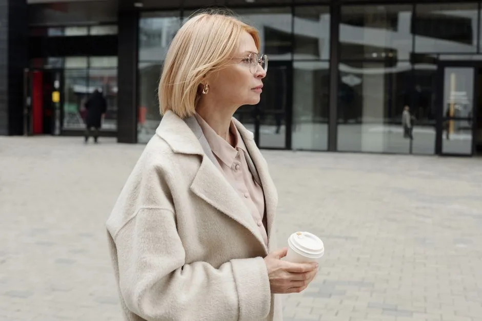 Woman in a beige coat holding coffee, standing confidently in an urban setting.
