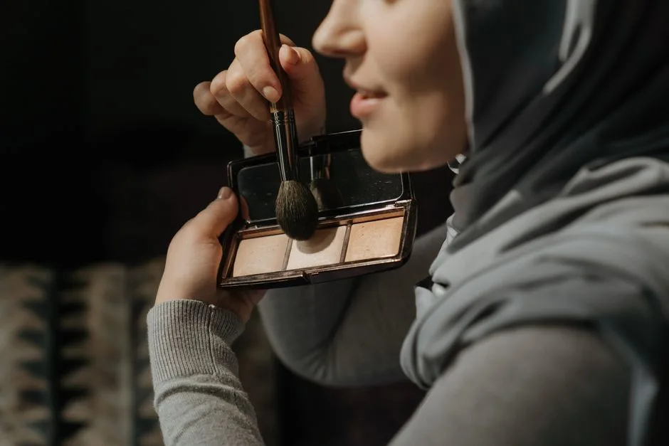 A close-up of a woman in a hijab applying makeup with a brush indoors.