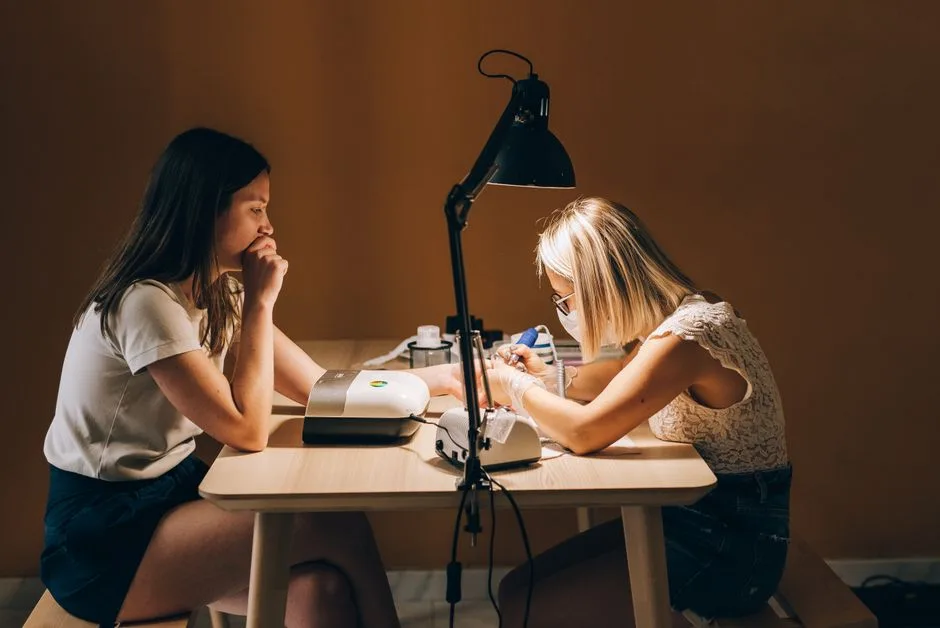 A manicurist wearing a face mask focuses on a customer's nails in a dimly lit room.
