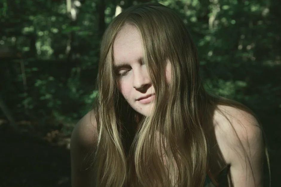 A blonde woman with closed eyes enjoys sunlight in a tranquil forest setting.