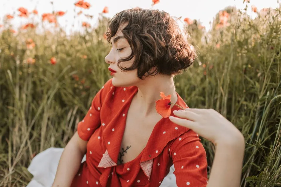 Brunette woman in red polka-dot dress posing in a flower field with red lips and short hair.