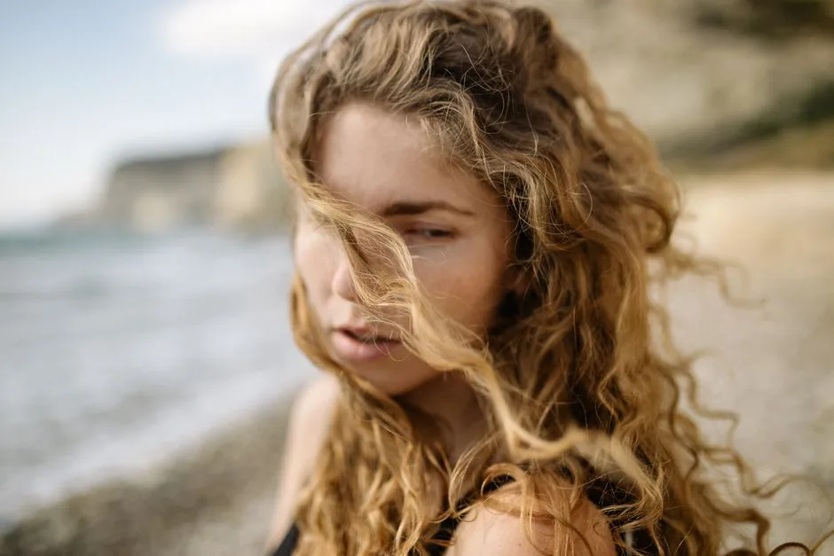 Young woman with curly blonde hair on a windy beach day.