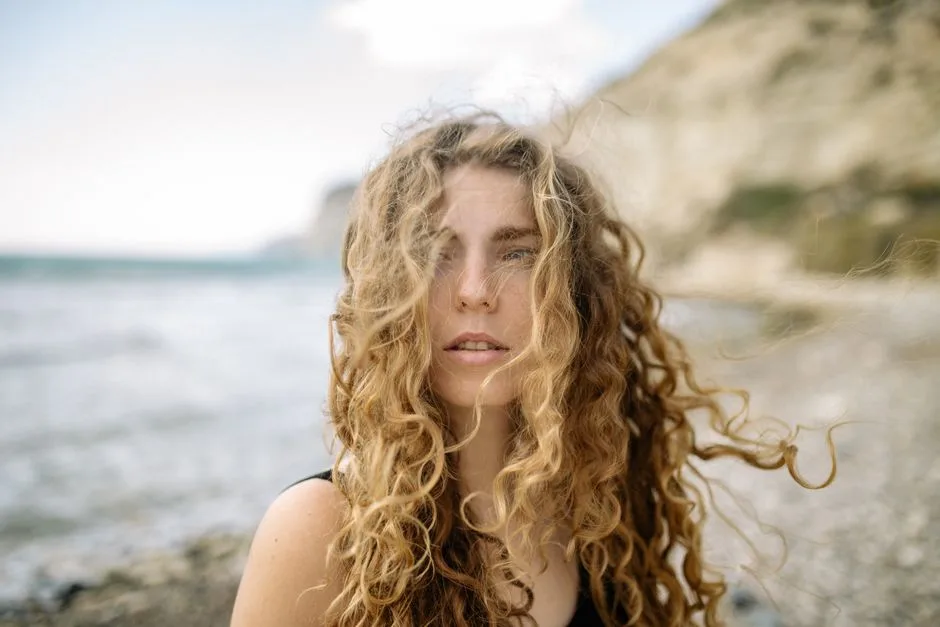 Close-up portrait of a woman with curly hair on a sandy beach with a blurred background.