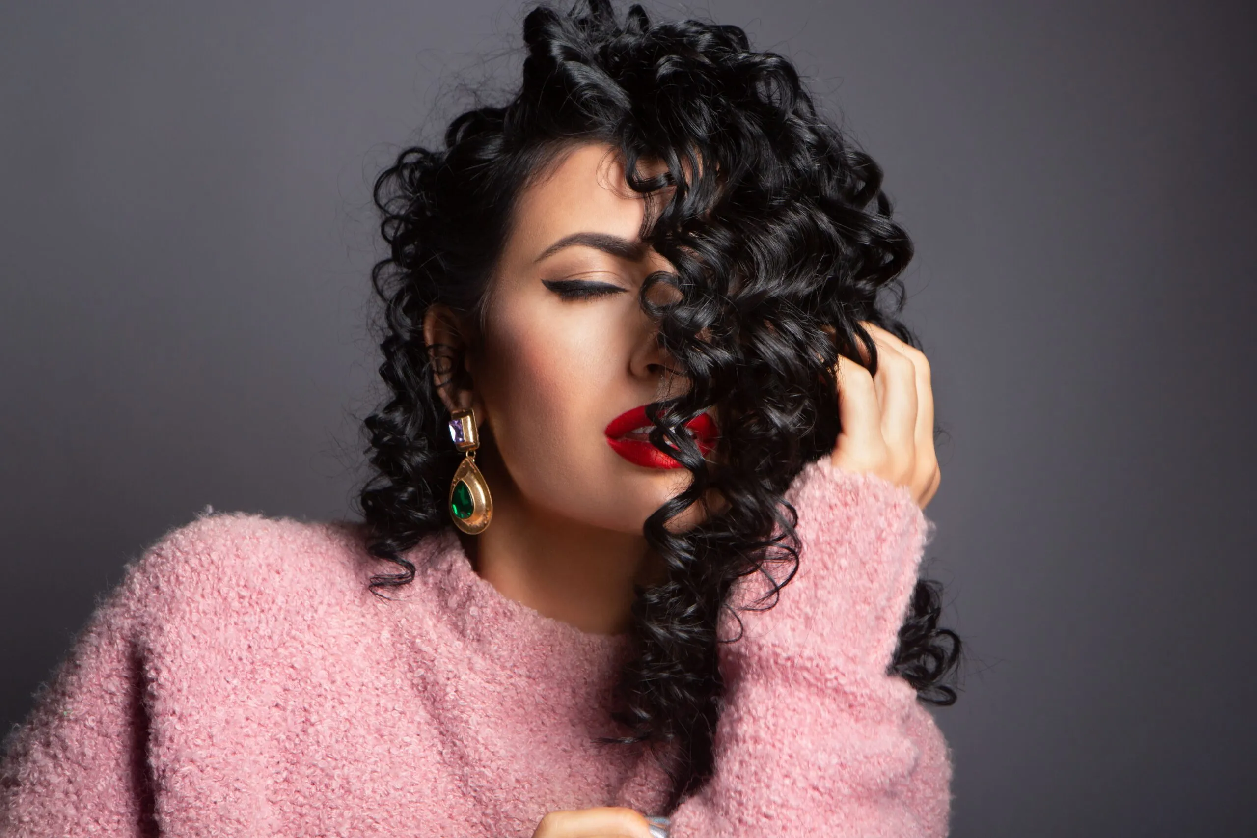 Sensual female with closed eyes and makeup touching curly hair gently against gray background