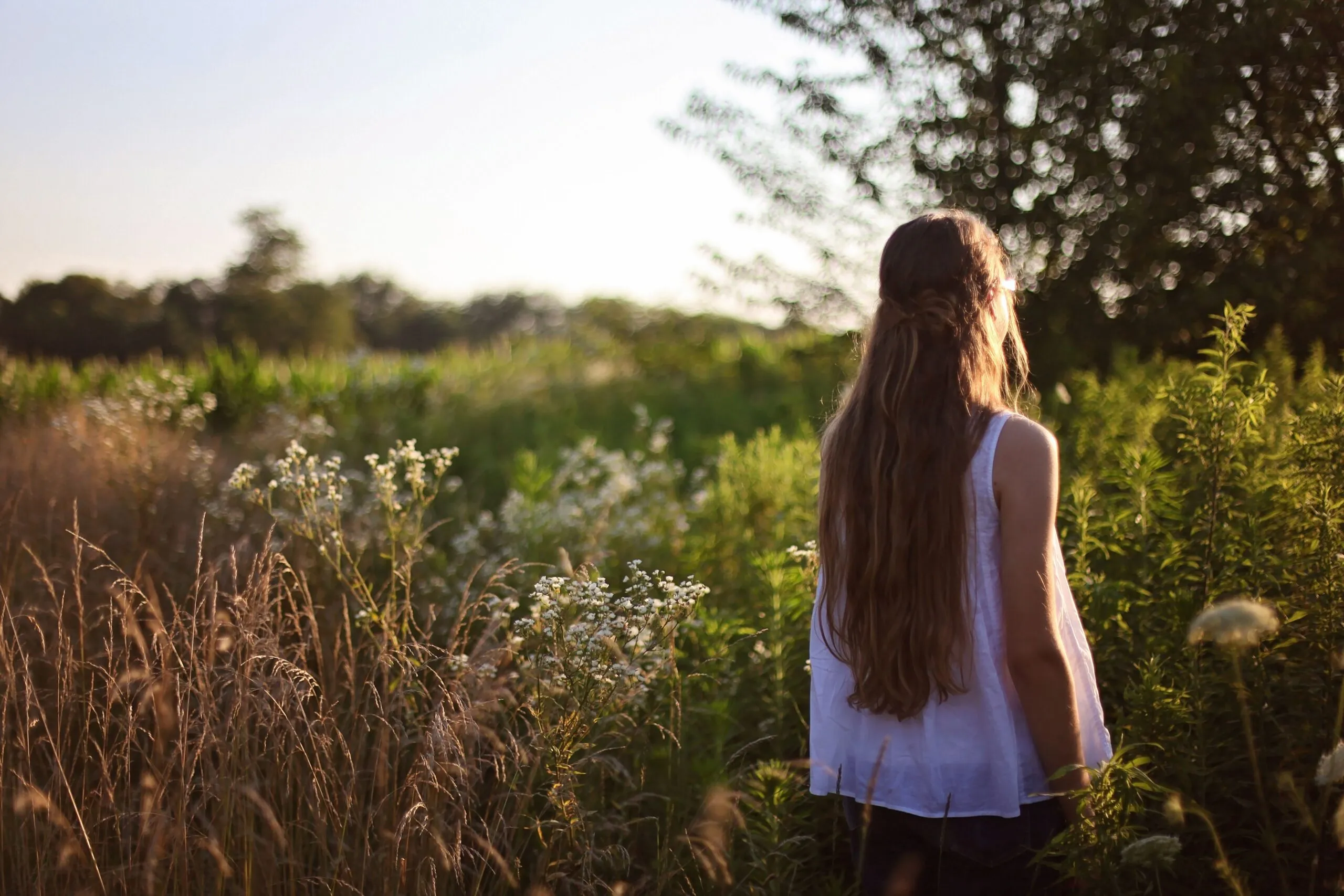 A woman with long hair stands in a sunlit countryside field surrounded by wildflowers.