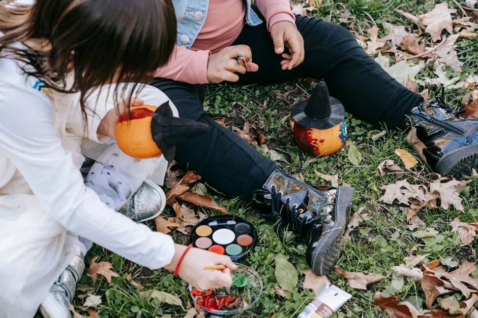 Crop unrecognizable little girls sitting on fallen leaves with paints and decorating pumpkins for Halloween
