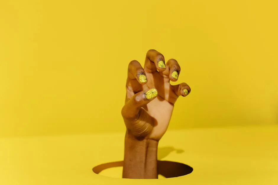 Close-up of a hand with yellow cracked nail art emerging through a yellow surface.