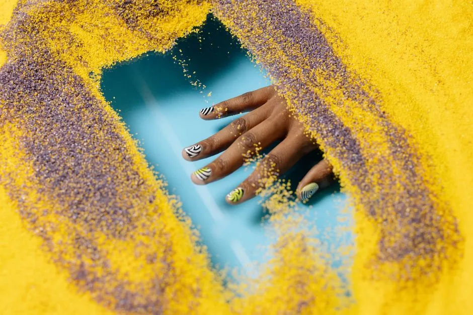 Stylish hand with creative nail art surrounded by colorful sand, showcasing vibrant design.