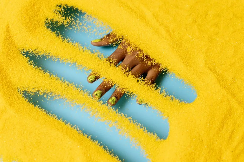 A striking image of a hand with yellow manicure submerged in vibrant yellow powder, showcasing creative nail art.