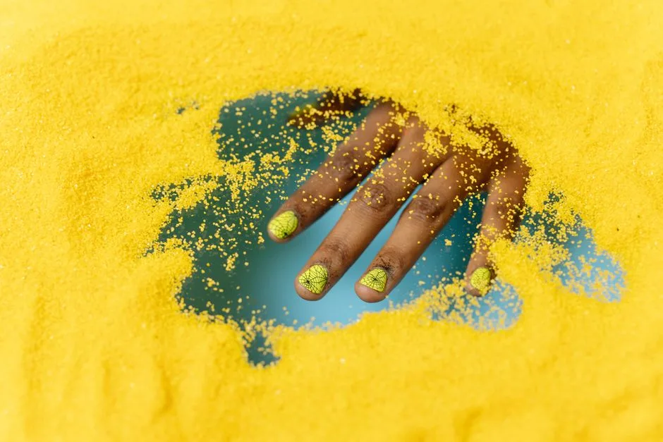 Close-up of a hand with creative nail art partially buried in vibrant yellow sand.