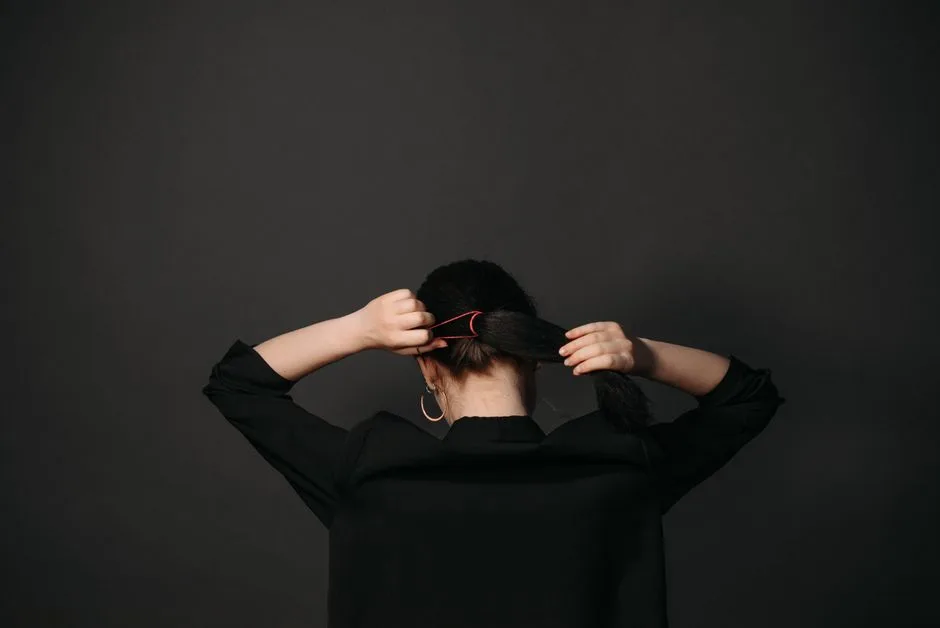 Woman tying her hair into a ponytail against a dark background, focusing on hairstyle and casual elegance.