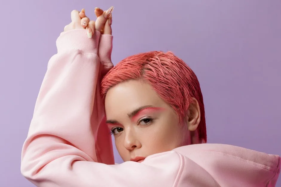 Close-up portrait of a young woman with pink hair and eye shadow against a violet background.