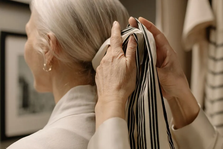 Elegant senior woman tying a striped scarf in her gray hair, showcasing style and grace.