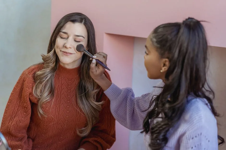 Side view of young ethnic woman applying foundation powder on face of smiling female friend sitting on chair with closed eyes