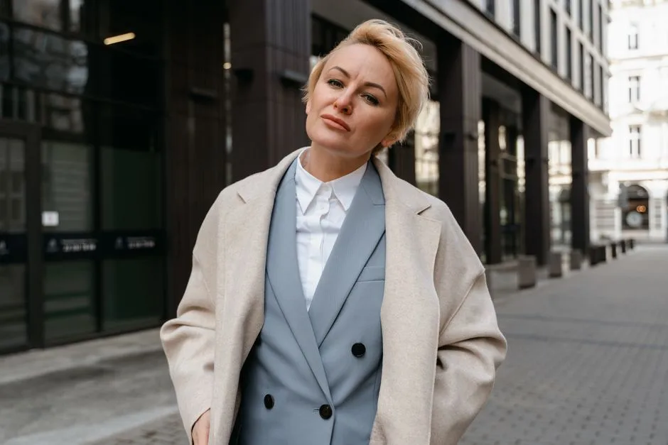 Confident woman in a blazer posing outdoors in a city with modern architecture.