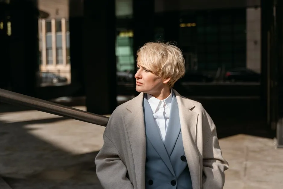 Professional blonde woman in a coat, facing away in a sunlit urban environment.