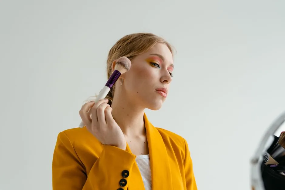 A woman in a yellow blazer applies makeup with a brush, showcasing a glamorous look.