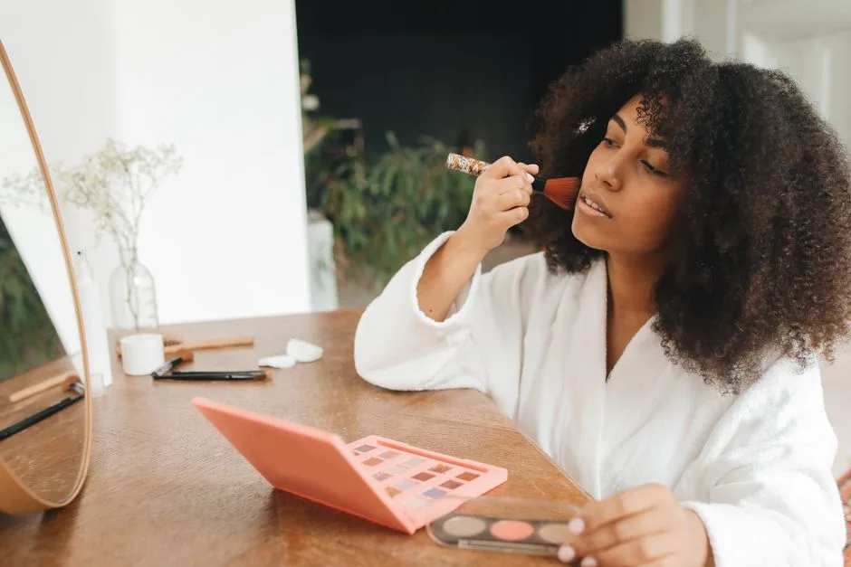A woman with curly hair applies makeup at home using a brush and mirror, seated at a wooden table.