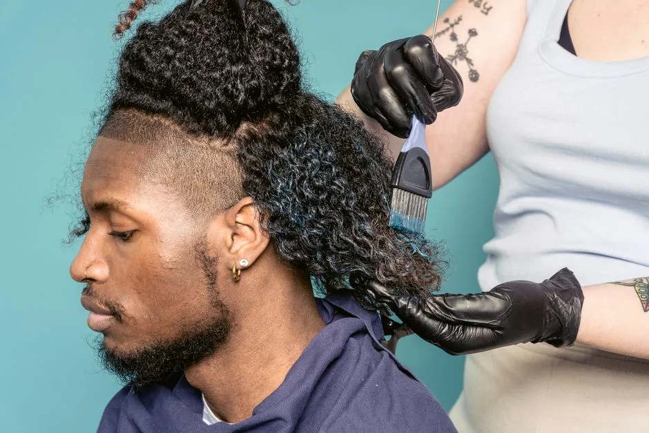 Crop female hairdresser in gloves with brush spreading dye on hair of African American male customer sitting against blue background