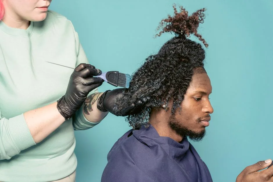 Crop hairstylist with tattoo dying African American male customer using cellphone against blue background