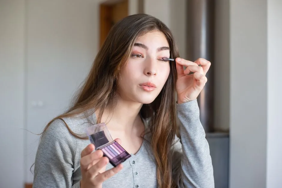 Asian woman applying makeup with eyeshadow palette indoors, showcasing beauty routine.