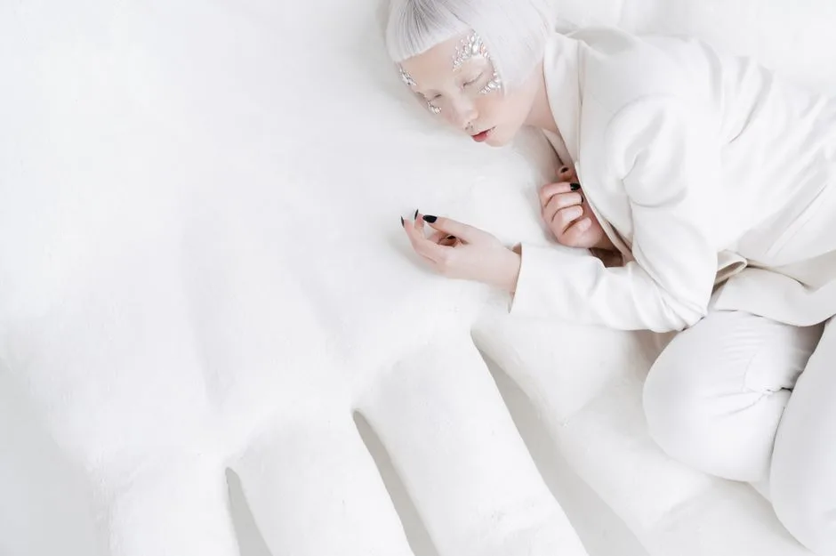 Stylish woman with closed eyes and artistic makeup in a white suit, lying down in a minimalist setting.