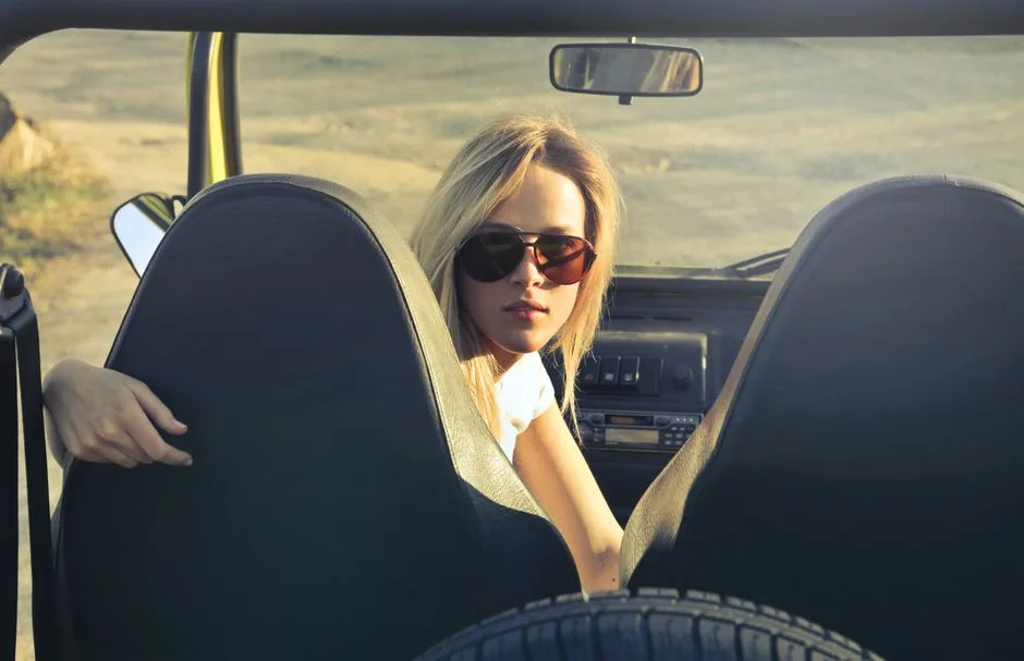 Blonde woman in sunglasses sitting in a convertible on a sunny day, enjoying an outdoor adventure.