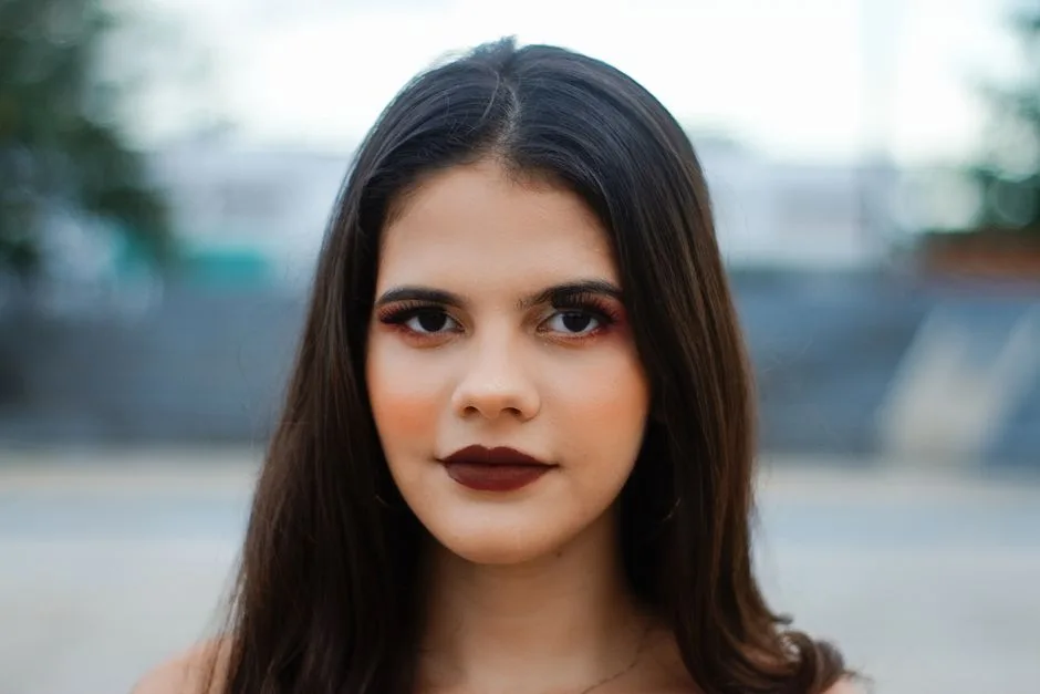 A close-up portrait of a woman with striking makeup, shot outdoors with a blurred background.