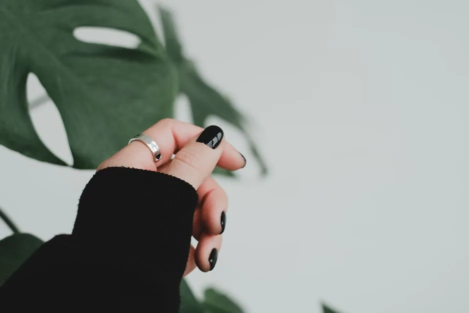 Close-up of a hand with black manicured nails and a silver ring against a green leaf background.