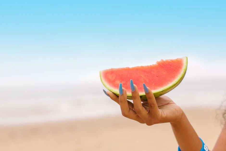 Vibrant watermelon slice held by a hand with blue nails on a sunny beach day.