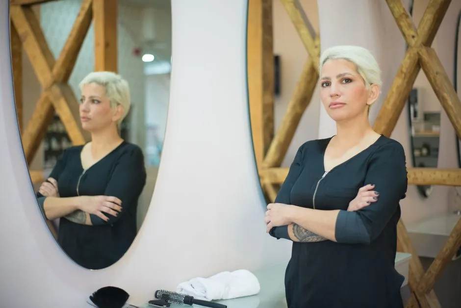 A professional hairdresser confidently stands in a modern salon with crossed arms, reflecting in a mirror.