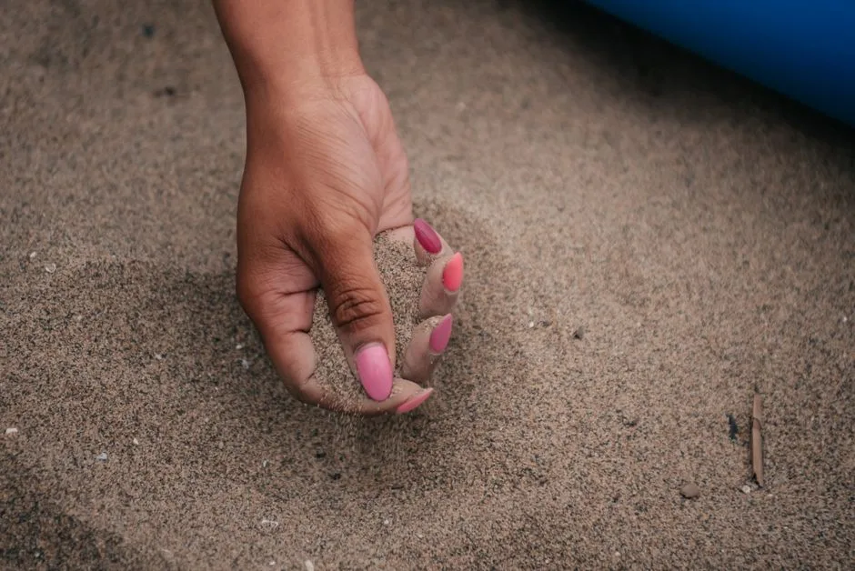 A close-up photo of a hand with pink manicured nails holding sand on a beach.