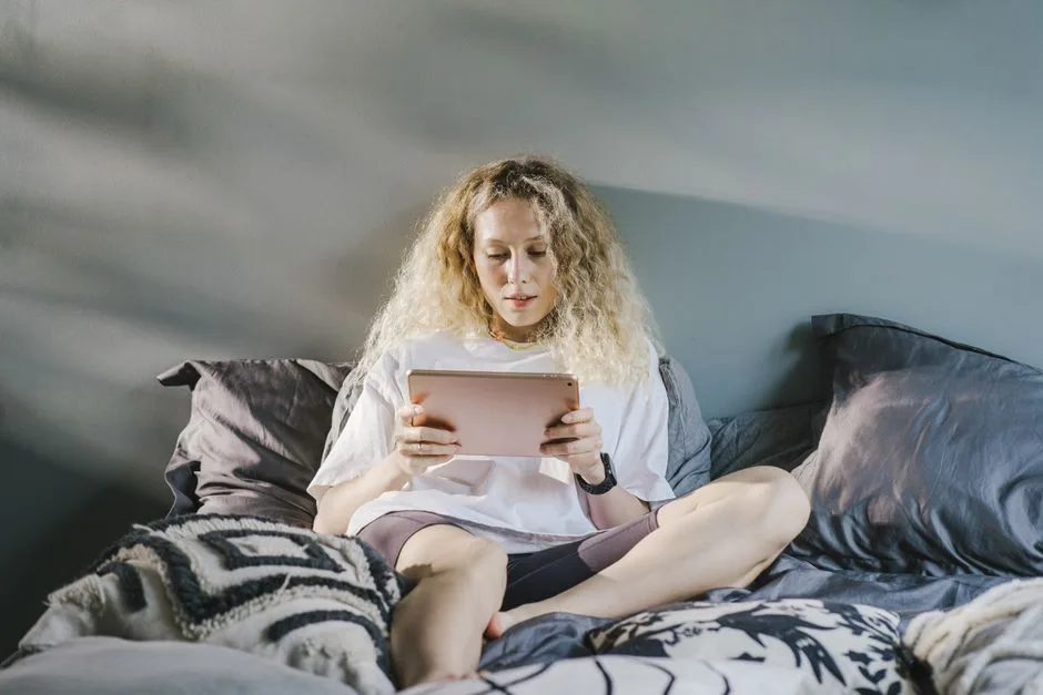 A woman with blonde hair sits on a cozy bed using a digital tablet indoors.