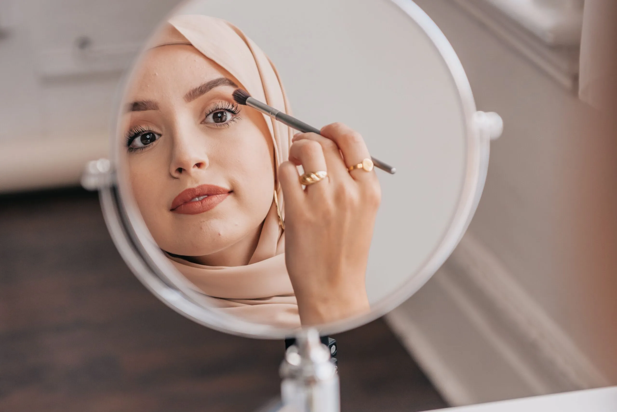 Close-up of a woman applying makeup with a brush, reflecting in a round mirror indoors.