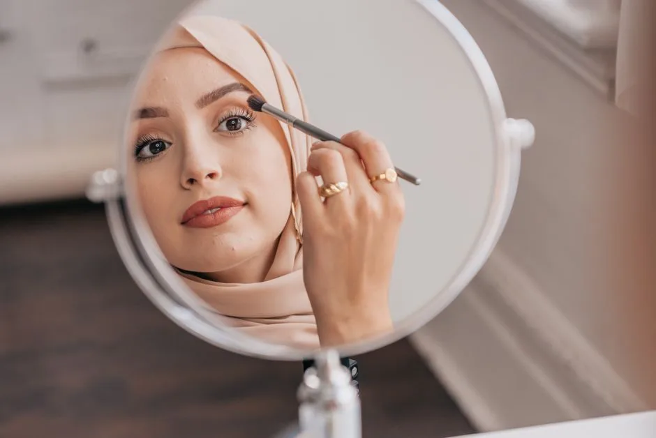 Close-up of a woman applying makeup with a brush, reflecting in a round mirror indoors.