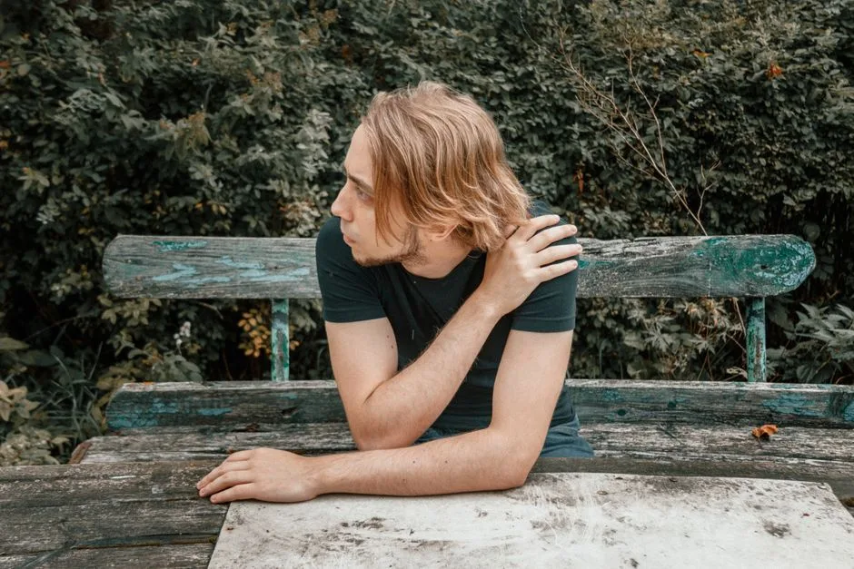 Contemplative young man with long blond hair sitting on a wooden bench outdoors.